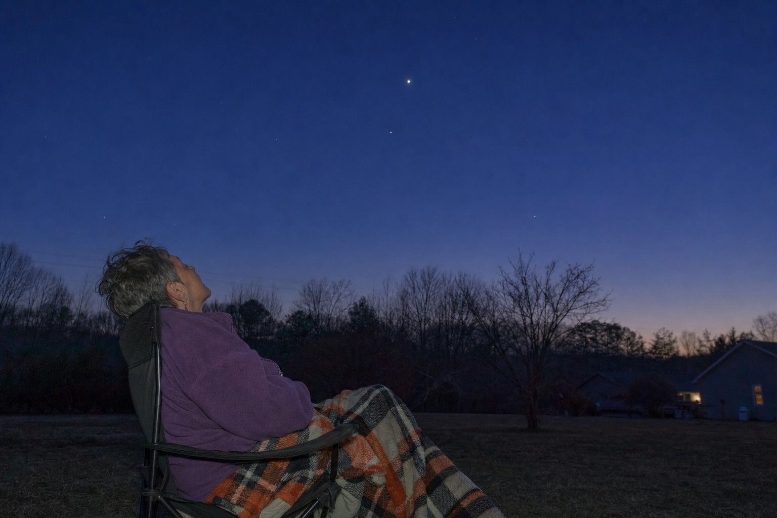 An older adult sits in a folding camp chair in a backyard at deep-blue dusk, head tipped back to look up at a darkening sky with a few early stars