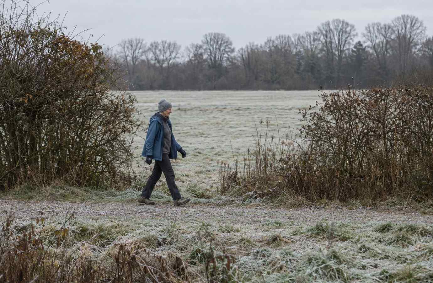 A lone walker in layered winter clothing crossing a frosted field path under flat grey light