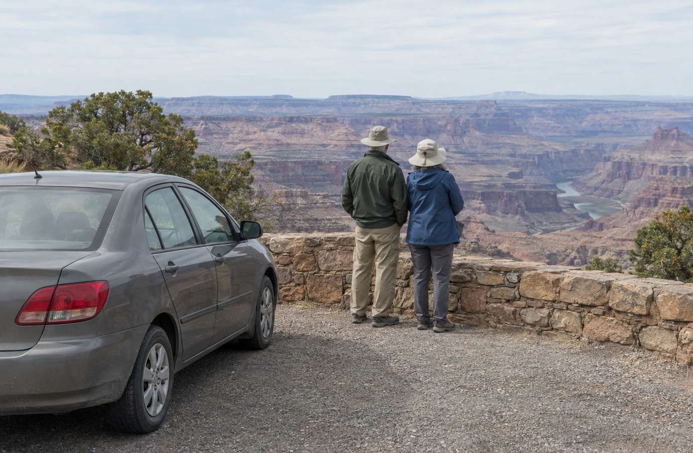 Two older travelers standing at a low stone wall beside a parked car, looking out over a wide canyon